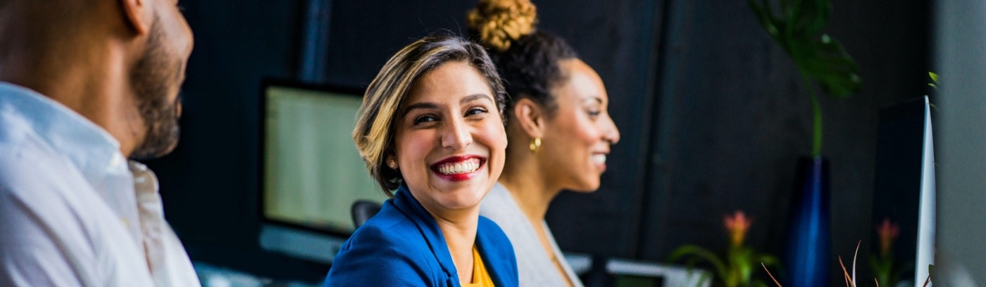 A woman smiles while seated at a desk, accompanied by two other individuals, fostering a friendly work environment.