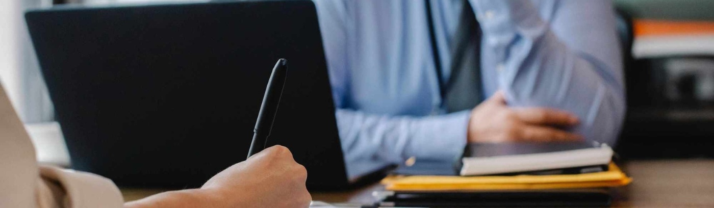 A seated individual works on a laptop at a desk, accompanied by another person holding a pen.