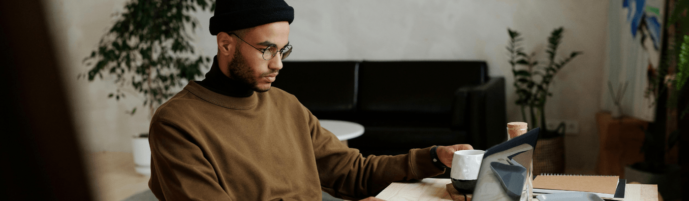 A man in a beanie and sweater is seated at a table, working on his laptop.