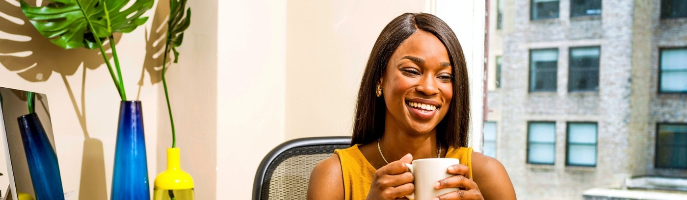 A woman with a cheerful smile sits at her desk, cradling a cup of coffee in her hands.