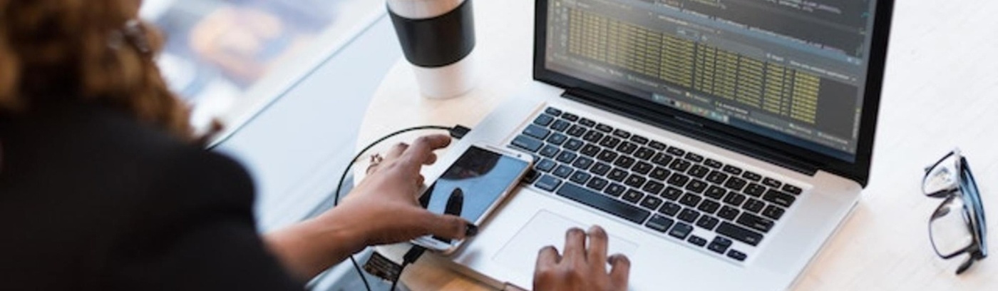 A person engaged with a laptop, simultaneously using a smartphone, seated at a workspace.