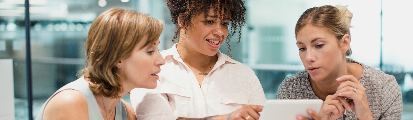 Three women in business attire collaboratively examining a tablet, showcasing teamwork and professionalism in a modern setting.