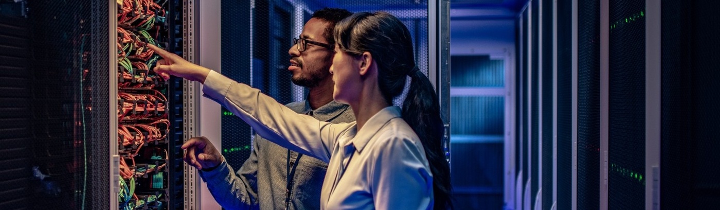 Two people in a server room with rows of servers and cables in the background.