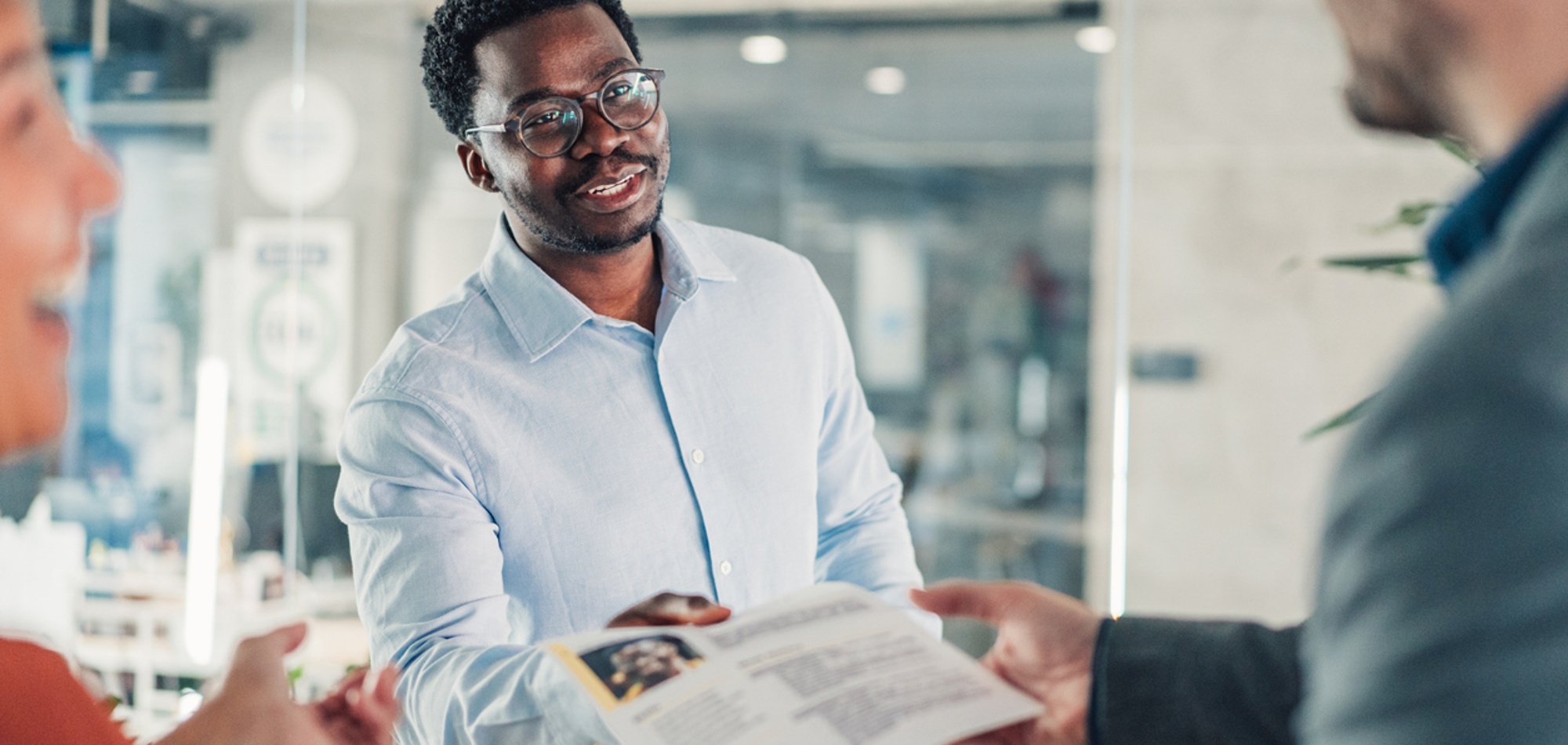 A professional man in a light blue shirt and glasses smiles while handing a document, possibly a resume or business proposal, to another person during an interview or meeting. The image captures a positive and professional interaction in an office setting.