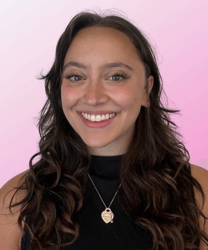 Woman with brown hair and a black top smiling in front of pink background