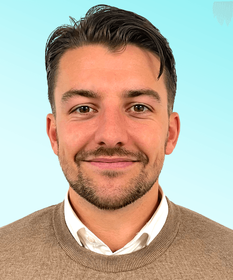 Man with brunette hair in collar shirt and knit jumper smiling in front of blue background