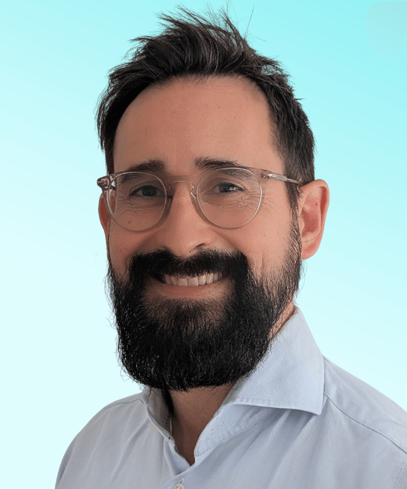 Man with dark hair and beard in a collar shirt in front of light blue background