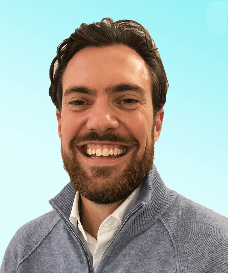 Man with dark hair and beard wearing a grey knit zip sweater smiling in front of blue background