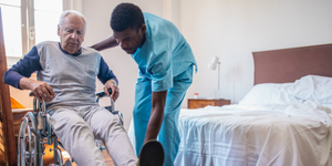 A Healthcare Assistant helping an elderly resident get out of their bed and into a wheelchair