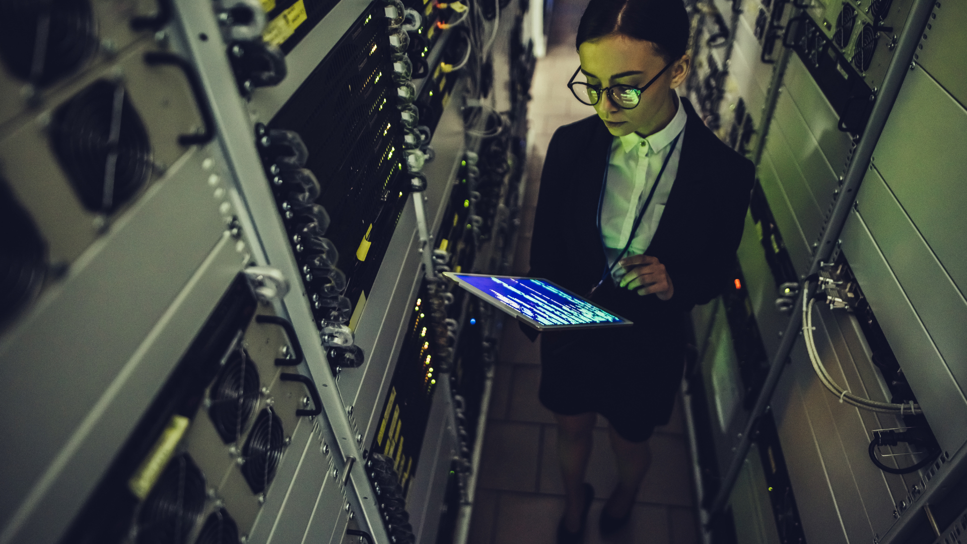 A professional woman in formal attire and glasses stands in a dimly lit server room, using a tablet displaying lines of code, surrounded by server racks with blinking lights and connected cables.