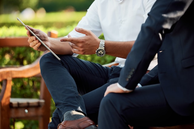 A close-up image of two men sitting on a park bench. One man, wearing a white shirt and black pants, holds a tablet and points to the screen while talking to the other man, who is wearing a dark suit.