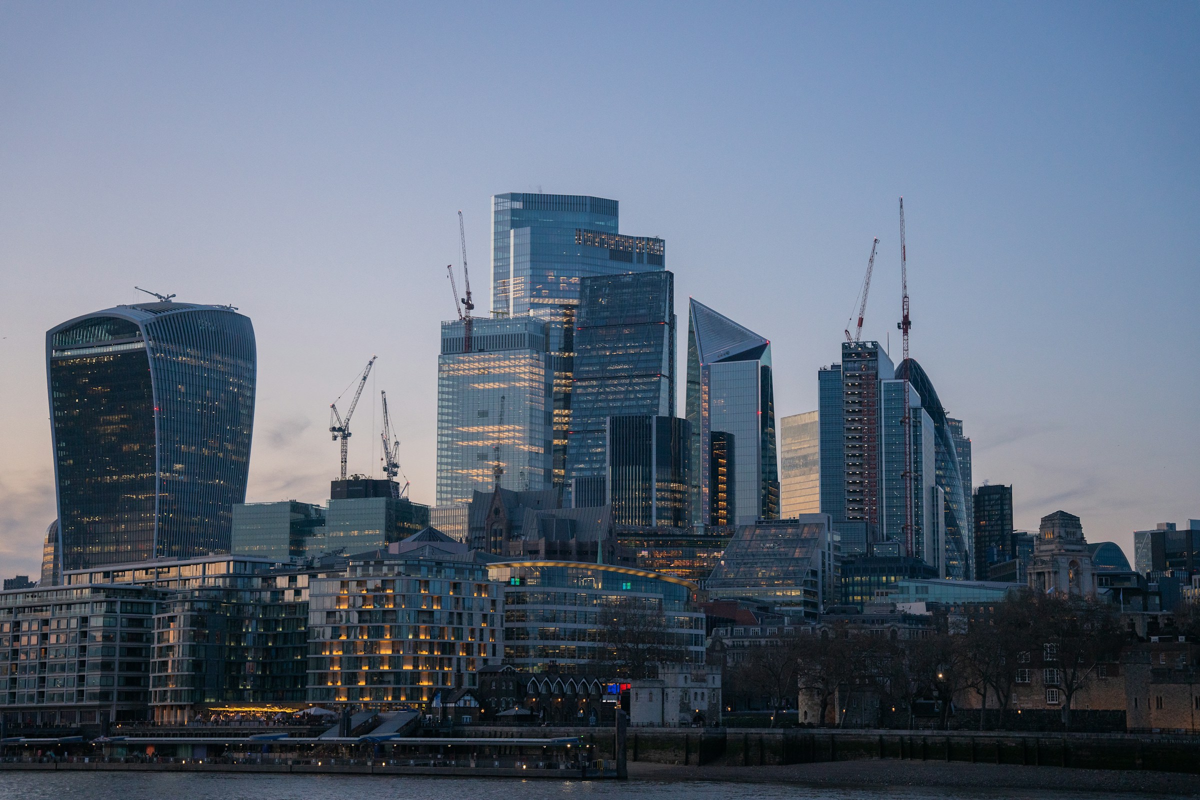 View of London’s modern financial district at dusk featuring iconic skyscrapers including the Walkie-Talkie building, with construction cranes visible and warm interior lights beginning to glow across glass façades.